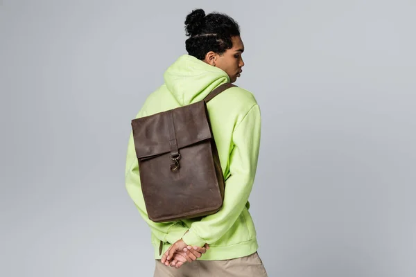 Hombre afroamericano con capucha verde que lleva la mochila de cuero aislada en gris — Stock Photo