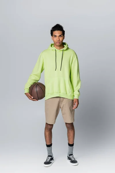 Vista completa del hombre afroamericano en sudadera con capucha beige y pantalones cortos verdes sosteniendo baloncesto sobre fondo gris - foto de stock