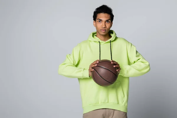 Afro americano cara no verde hoodie olhando para a câmera enquanto segurando basquete isolado no cinza — Fotografia de Stock