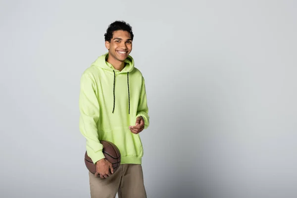 Élégant homme afro-américain en sweat à capuche vert souriant à la caméra tout en se tenant debout avec basket sur fond gris — Photo de stock