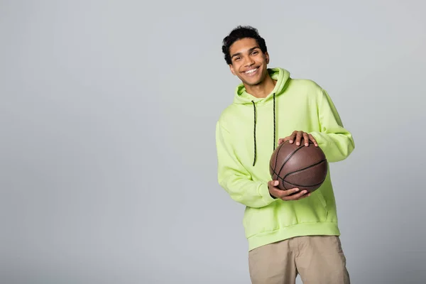 Fröhlicher afrikanisch-amerikanischer Mann mit Basketballkorb, während er isoliert auf grau in die Kamera lächelt — Stockfoto