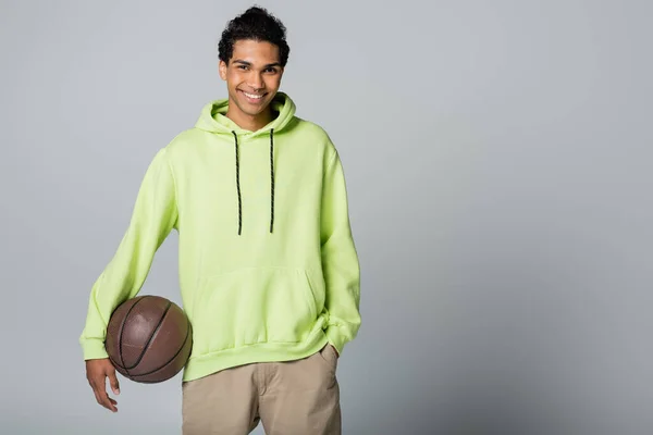 Joven afroamericano con capucha verde y pantalones cortos beige de pie con baloncesto sobre fondo gris - foto de stock