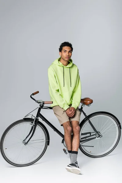 Joven afroamericano hombre en verde sudadera con capucha mirando a la cámara cerca de la bicicleta sobre fondo gris - foto de stock
