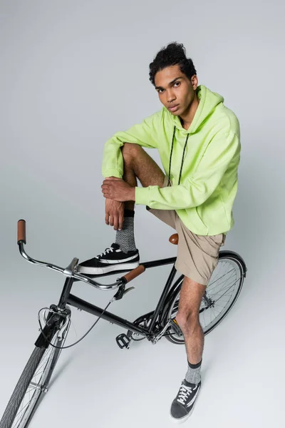 Hombre afroamericano de moda mirando la cámara cerca de la bicicleta sobre fondo gris - foto de stock