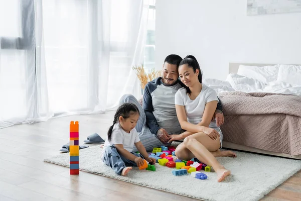 Cheerful asian parents looking at toddler daughter playing building blocks — Stock Photo