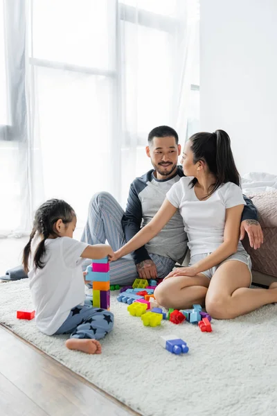 Cheerful asian husband and wife looking at each other while toddler daughter playing building blocks — Stock Photo