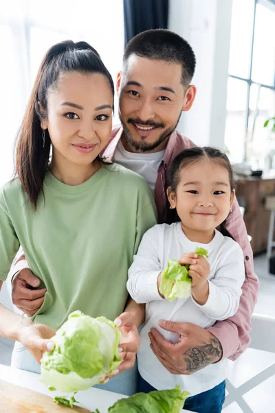 Щасливий азіатський чоловік і жінка дивиться на камеру і посміхається біля дочки зі свіжим салатом — стокове фото