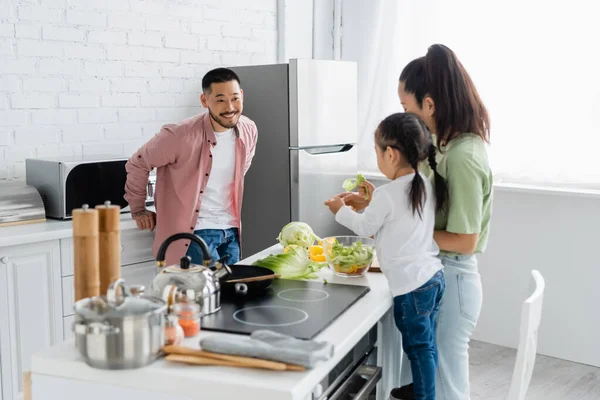 Feliz asiático hombre mirando borrosa esposa y hija preparando ensalada en cocina - foto de stock