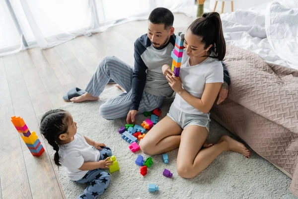 Alto ángulo vista de asiático marido y esposa mirando a niño pequeño hija jugando bloques de construcción - foto de stock