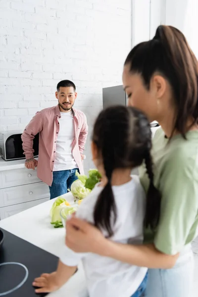 Усміхнений азіатський чоловік дивиться на розмиту дружину з дочкою на кухні — стокове фото