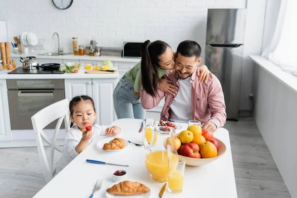 Asiatique femme baisers mari près de fille avoir petit déjeuner dans cuisine — Photo de stock