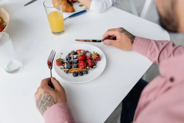 Vista cortada de homem tatuado segurando talheres perto de waffles com bagas na placa — Fotografia de Stock