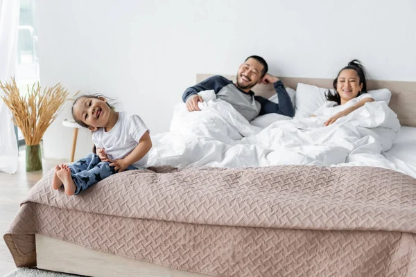 Cheerful asian kid sitting on bed near blurred parents — Stock Photo