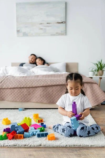 Toddler kid playing building blocks near blurred parents lying on bed on background — Stock Photo