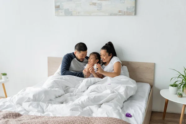 Mãe asiática segurando blocos de construção perto alegre criança e marido na cama — Fotografia de Stock