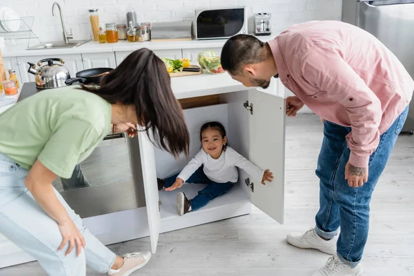 Happy toddler asian kid hiding in kitchen cabinet near parents — Stock Photo