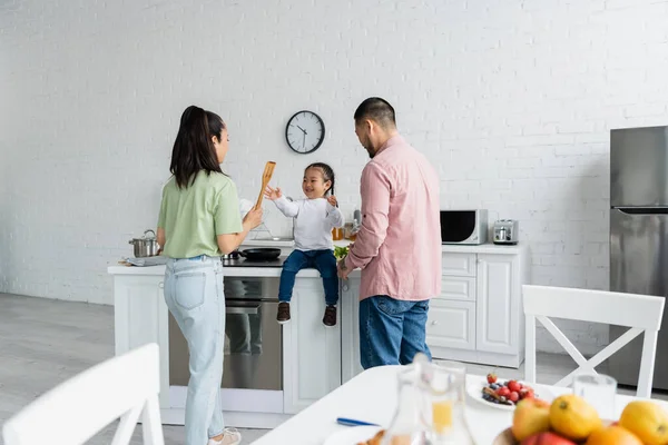 Happy asian kid reaching wooden spatula in hand of mother while sitting near dad — Stock Photo