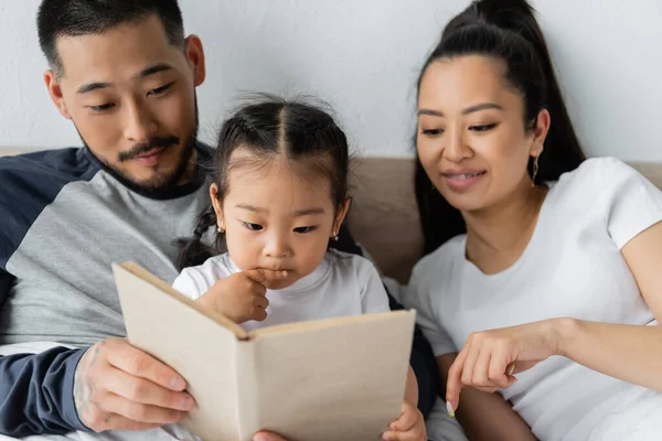 Happy asian parents reading book to toddler daughter in bed — Stock Photo