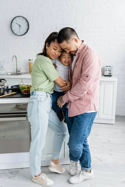 Asian parents hugging with cheerful toddler daughter in kitchen — Stock Photo