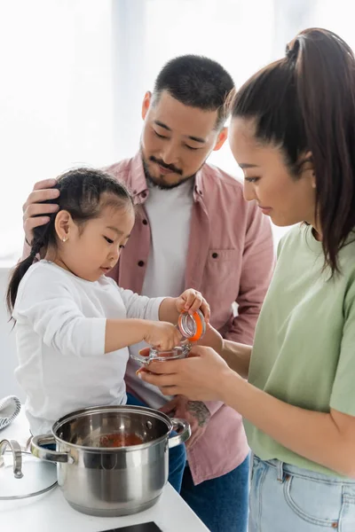 Asiático niño llegar botella con especias cerca padres mientras cocinar en cocina - foto de stock