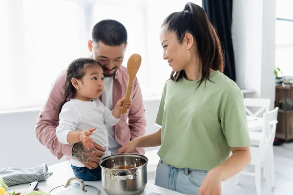 Asiatische Mutter lächelt, während sie ihre Tochter in der Nähe ihres Mannes in der Küche ansieht — Stockfoto