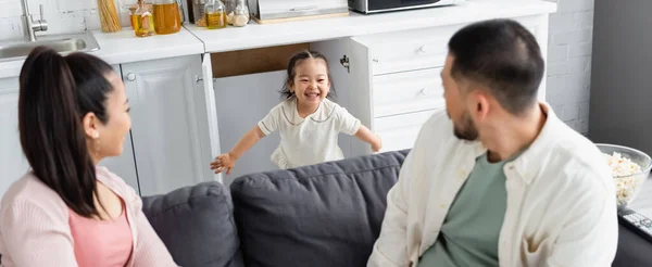 Feliz asiático niño escondido en la cocina gabinete cerca borrosa padres en sofá, bandera - foto de stock