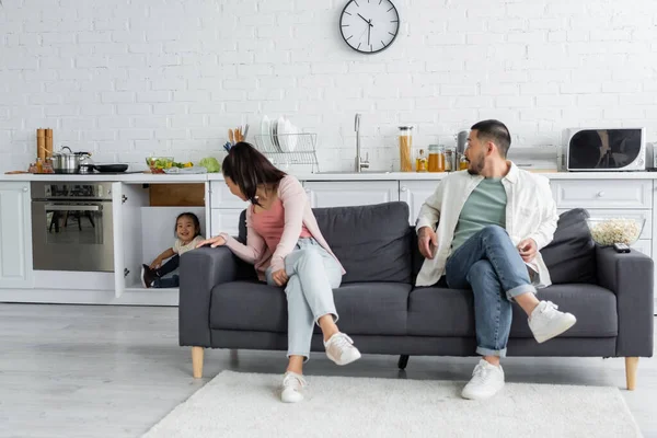 Happy asian kid hiding in kitchen cabinet near parents on sofa — Stock Photo