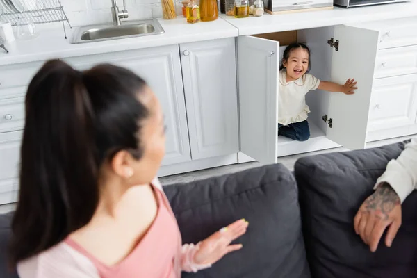 Cheerful asian kid hiding in kitchen cabinet near blurred parents on sofa — Stock Photo