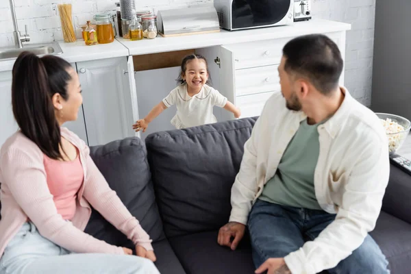 Joyful asian kid hiding in kitchen cabinet near blurred parents on sofa — Stock Photo