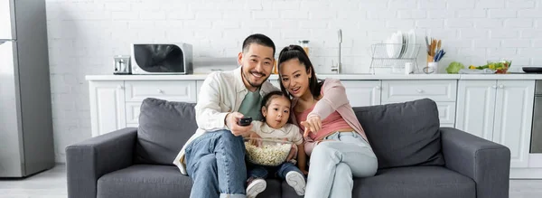 Freudige asiatische Familie schaut Film in der Nähe von Popcorn im Wohnzimmer, Banner — Stockfoto