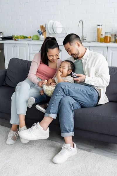 Feliz asiático padres sentado en sofá y mirando a hija comer palomitas de maíz - foto de stock