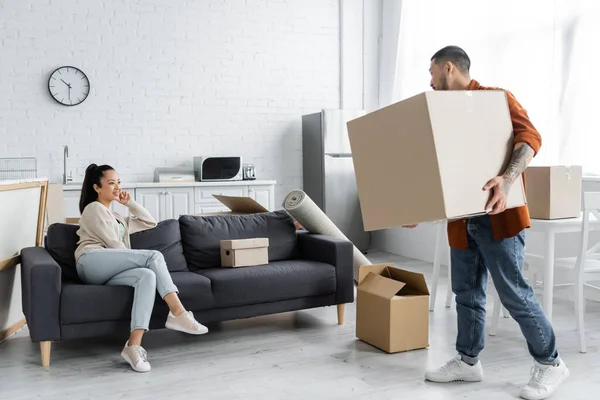 Asian man holding carton box and looking at wife sitting on couch — Stock Photo