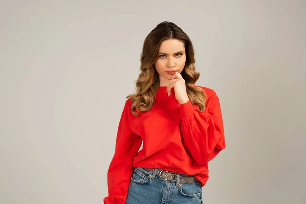 Thoughtful curly woman in red sweatshirt looking at camera isolated on grey — Stock Photo