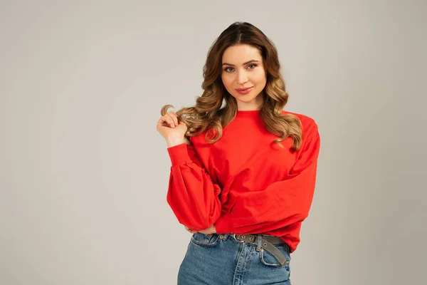 Femme heureuse en sweat-shirt ajustant les cheveux et regardant la caméra isolée sur gris — Photo de stock