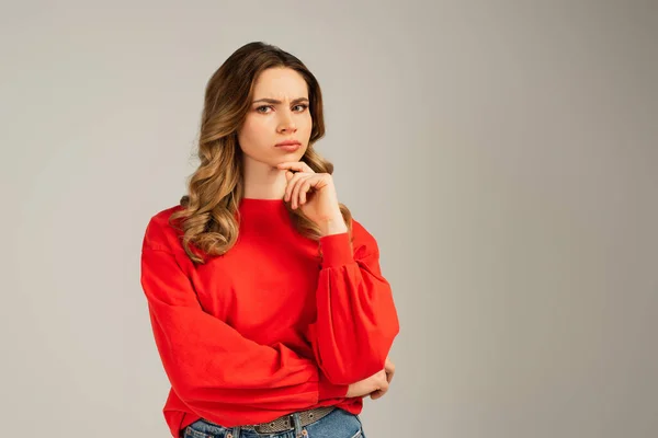 Pensive woman in red sweatshirt looking at camera and thinking isolated on grey — Stock Photo