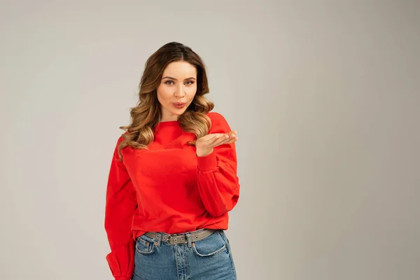 Frau in rotem Sweatshirt blickt in die Kamera und sendet Luftkuss isoliert auf grau — Stockfoto