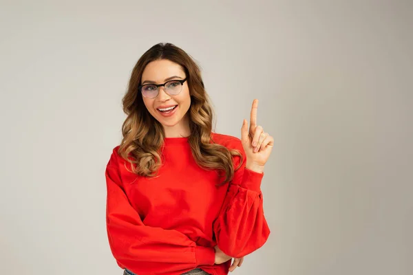 Cheerful woman in sweatshirt and eyeglasses pointing with finger isolated on grey — Stock Photo