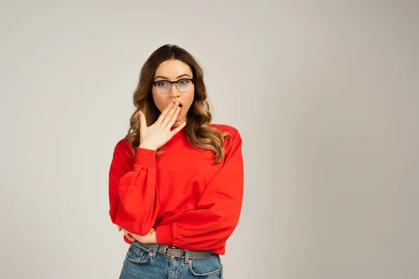 Schockierte Frau in Sweatshirt und Brille, die den Mund isoliert auf grau verdeckt — Stockfoto