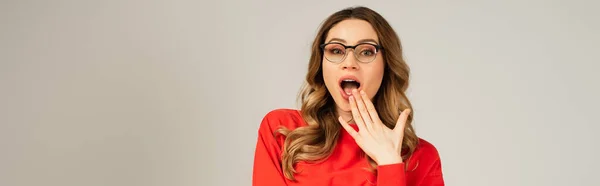 Shocked woman in sweatshirt and eyeglasses with open mouth isolated on grey, banner — Stock Photo