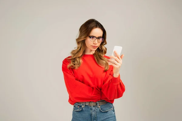 Femme mécontente dans les lunettes en utilisant smartphone isolé sur gris — Photo de stock