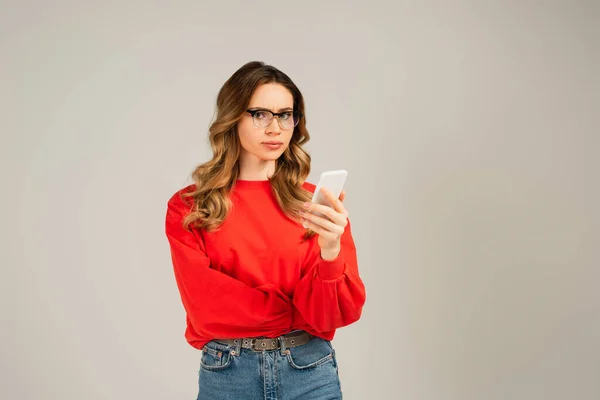 Кудрявая женщина в очках с помощью смартфона, изолированного на сером — стоковое фото