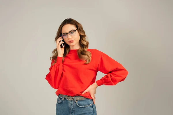 Mujer disgustada en gafas de vista hablando en smartphone mientras está de pie con la mano en la cadera aislada en gris - foto de stock