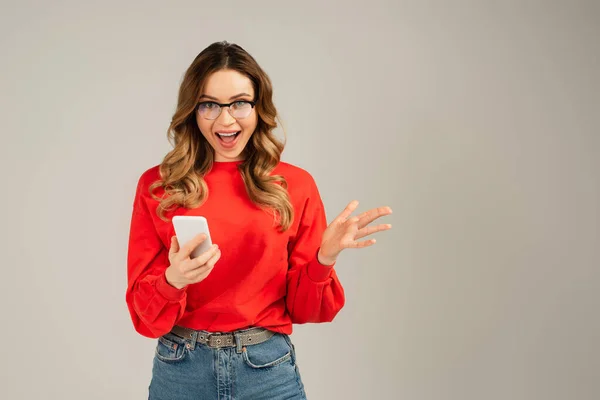 Збуджена жінка в окулярах тримає мобільний телефон ізольовано на сірому — стокове фото