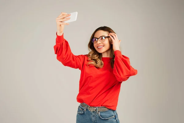 Mujer feliz en gafas y sudadera tomando selfie en smartphone aislado en gris - foto de stock