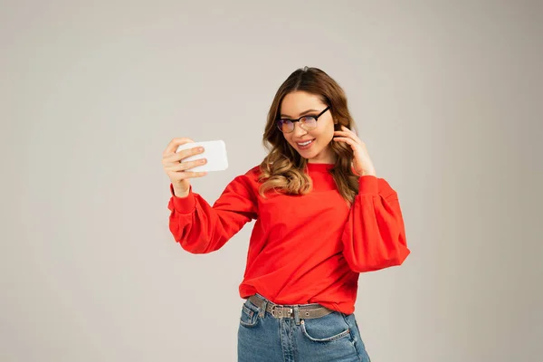 Положительная женщина в очках делает селфи на смартфоне, изолированном от серого — стоковое фото
