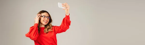 Happy woman in eyeglasses taking selfie on smartphone isolated on grey, banner — Stock Photo
