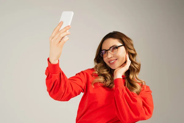 Mujer alegre en gafas tomando selfie en teléfono inteligente aislado en gris - foto de stock