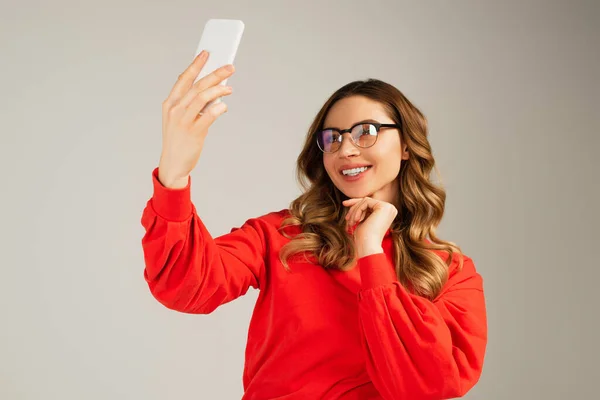 Счастливая женщина в очках делает селфи на смартфоне, изолированном от серого — стоковое фото