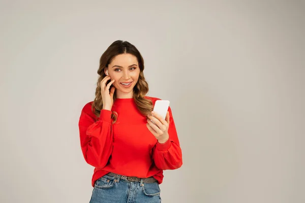 Glückliche Frau stellt drahtlose Kopfhörer ein, während sie Smartphone isoliert auf grau verwendet — Stockfoto