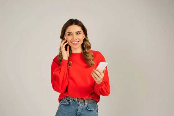 Cheerful woman adjusting wireless earphones while using smartphone isolated on grey — Stock Photo